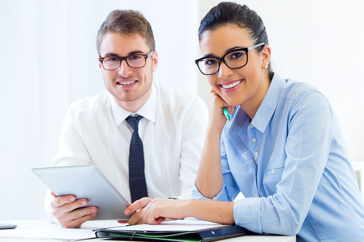 Two professionals similing, both wearing glasses in a business environment while the man holds a tablet