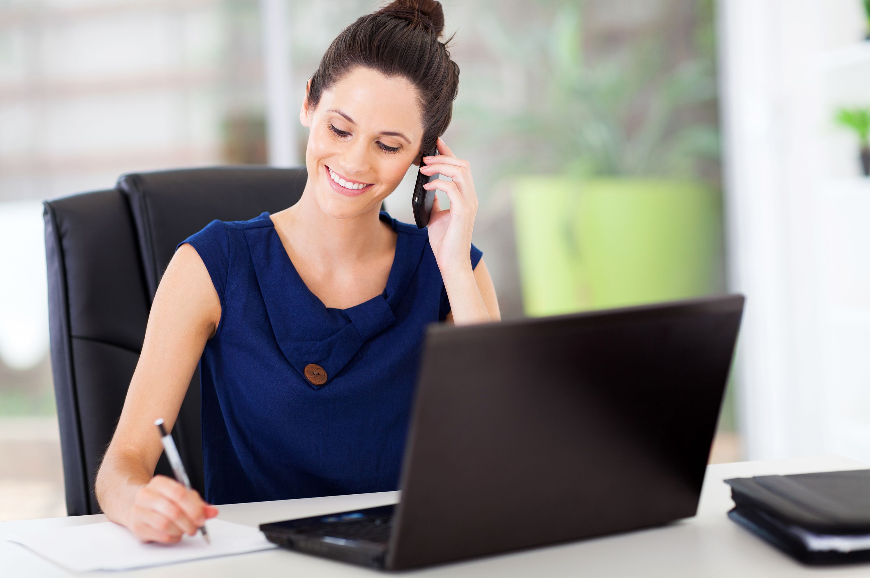 Business woman multitasking with laptop, writing on paper, and talking on phone with a smile