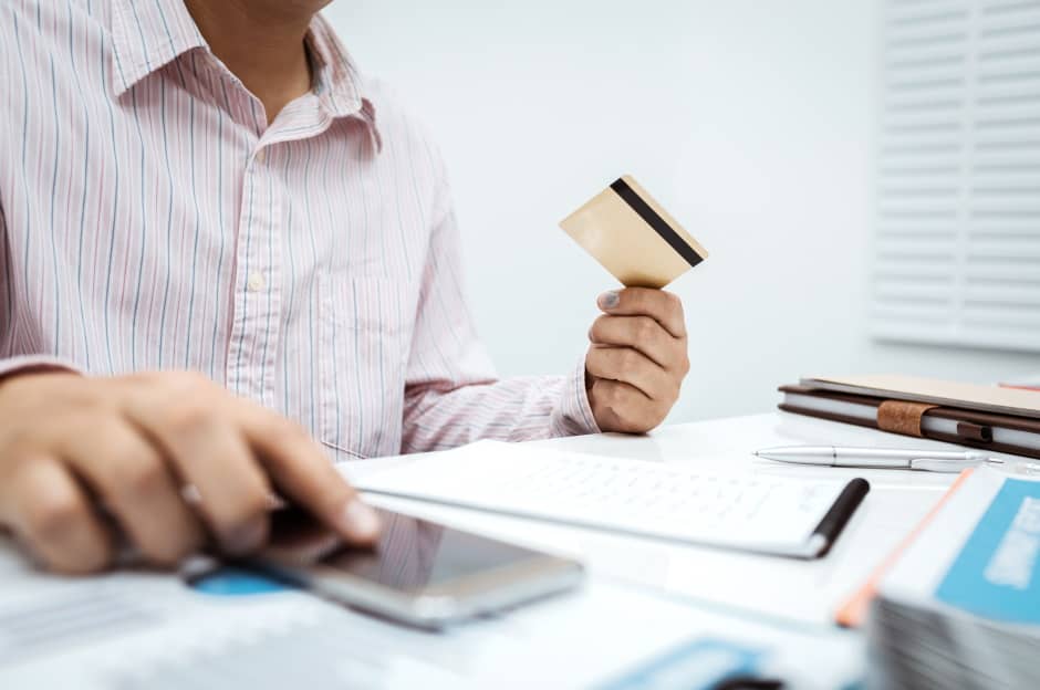 Man holding a card in one hand and using a cellphone in the other hand on a table with paperwork.