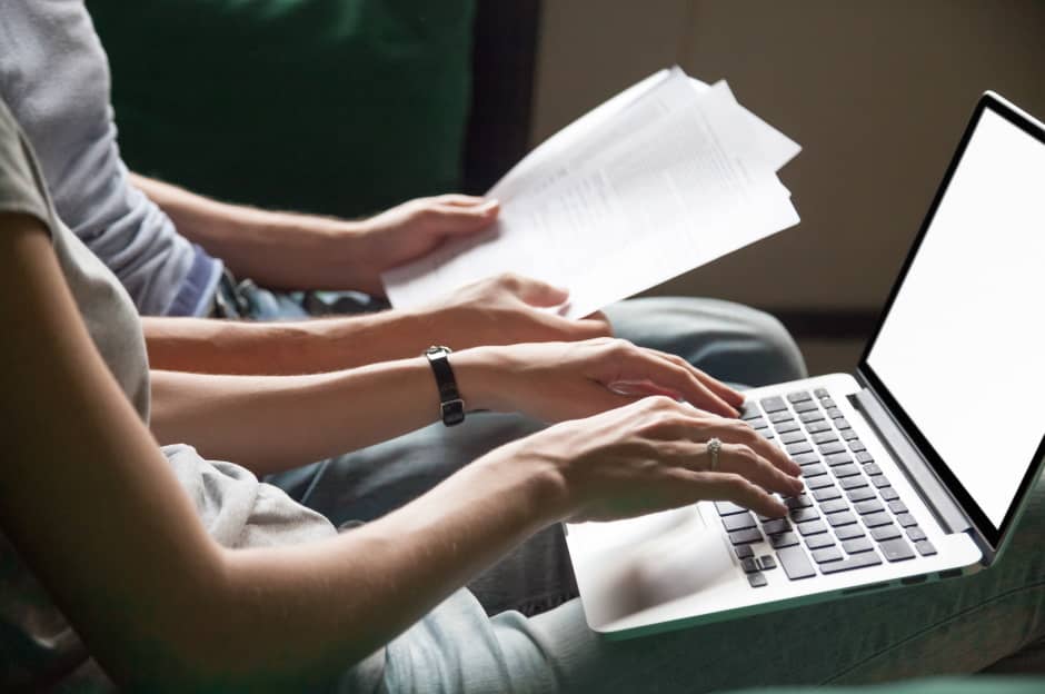 Women typing on a laptop while sitting with a man holding papers beside her